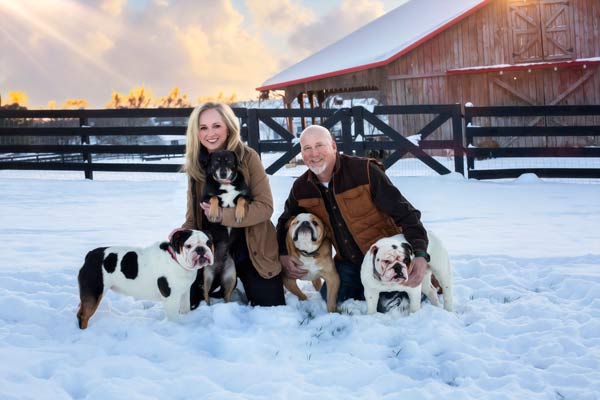 A couple sitting the snow in front of their barn surrounded by their 4 dogs